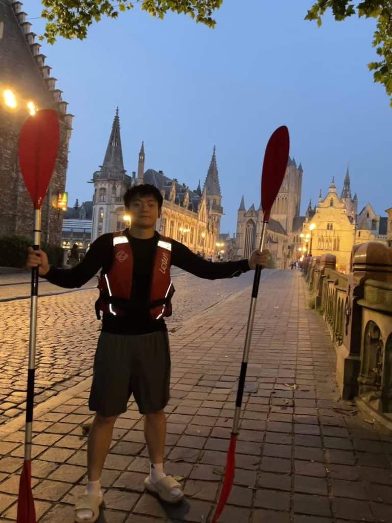 Ju Lin on the cobbled quay in Ghent at dusk holding two kayak paddles, the lit spires of the old town behind