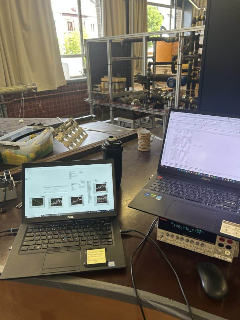 Two laptops on the lab bench, one running the LabVIEW acquisition program with live measurement plots, a bench multimeter beside them and the test rig behind